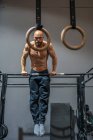 Full body shirtless bearded sportsman hanging on bar and doing pull ups during intense training in contemporary gym looking at camera — Stock Photo