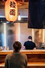 Back view of faceless black haired woman in sweater sitting at counter in cozy ramen bar — Stock Photo