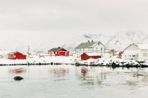 Холодное море со спокойной водой, расположенное рядом с прибрежным поселением и снежным горным хребтом в пасмурный зимний день на Лофских островах, Норвегия — стоковое фото