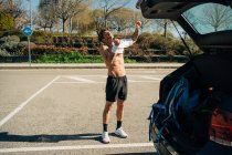 Männlicher Sportler mit Tätowierung streift an sonnigem Tag T-Shirt gegen Auto auf Parkplatz — Stockfoto