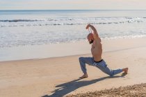 Vista lateral del atleta masculino sin camisa estirando los brazos con banda elástica mientras hace un salón haciendo ejercicio en la playa soleada vacía - foto de stock