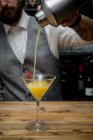 Crop unrecognizable barman pouring alcohol orange cocktail from shaker in glass placed on wooden counter in bar — Stock Photo