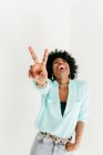 Verspielte junge Afroamerikanerin im trendigen Outfit hat Spaß und zeigt Friedenszeichen auf weißem Hintergrund — Stockfoto