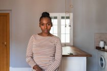 Young African American female with hair bun looking at camera against table and washing machine in kitchen — Stock Photo