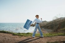 Side view of positive young female artist in stylish outfit and hat walking near sandy beach of wavy sea with painting in hand — Stock Photo