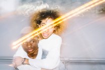 Alegre jovem afro-americano masculino dando passeio de piggyback para namorada feliz com cabelo encaracolado na roupa da moda perto de luzes congeladas — Fotografia de Stock