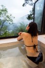 Relaxed young ethnic lady in swimwear sitting in Japanese onsen bath in spa resort next to window with view of mountains and green trees — Stock Photo