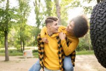 Young man giving piggyback ride to optimistic black girlfriend casual clothes while standing near car on sunny day in countryside — Stock Photo