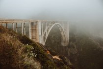 Захватывающий дух пейзаж удивительного белого арочного моста Биксби через реку в глубокой долине против склонов гор, покрытых пышной зеленой травой и полевыми цветами в Биг-Сур в США — стоковое фото