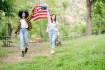 Веселая лесбийская многорасовая пара женщин бегает с национальным американским флагом по тропинке в лесу и улыбается, глядя в камеру — стоковое фото