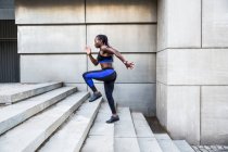 Vista lateral da mulher afro-americana forte correndo escadas enquanto treina perto do edifício moderno na rua da cidade — Fotografia de Stock