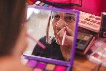 Junge Frau blickt in Spiegel und malt Ornament ins Gesicht, während sie sich im Atelier kreativ schminkt — Stockfoto