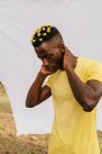 Beau mâle afro-américain avec des fleurs jaunes dans les cheveux touchant le cou et regardant vers le bas sur fond blanc — Photo de stock