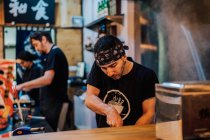 Young man in apron cooking Asian dishes while standing at counter in ramen bar — Stock Photo