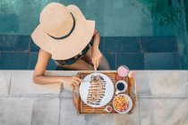 Сверху анонимная туристка в соломенной шляпе сидит в бассейне и режет вкусные блины с шоколадным соусом — стоковое фото