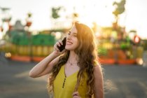 Conteúdo feminino falando no celular enquanto está de pé no parque de diversões à noite no verão — Fotografia de Stock