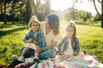 Feliz madre joven con lindas hijas con ropa similar disfrutando de un soleado día de verano juntas mientras están sentadas en una manta en un césped cubierto de hierba en el parque - foto de stock