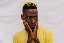 Handsome African American male with yellow flowers in hair touching face and looking away on white background — Stock Photo