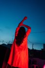 Rückansicht einer nicht wiedererkennbaren ethnischen Frau in Dessous, die in die Kamera schaut, während sie nachts auf der Terrasse unter rotem Neonlicht mit blauem dunklen Himmel ruht — Stockfoto