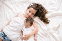 Vista dall'alto di allegra bambina che abbraccia adorabile neonato mentre giace su un letto morbido a casa — Foto stock