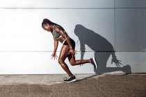 Muscular African American female in sportswear looking at camera and jumping while working out on city street near modern building wall — Stock Photo