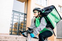 Low angle of female courier with thermal bag smiling and riding bike on street while making delivery on sunny day in city — Stock Photo