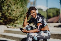 Junge Afroamerikanerin in bunter Sportbekleidung hört Musik über Kopfhörer, während sie im Park auf Steintreppen sitzt — Stockfoto