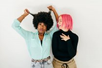 Cheerful young pink haired woman and African American girlfriend in stylish outfit standing together on white background — Stock Photo
