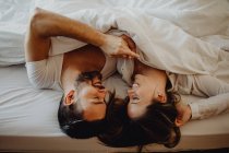 Веселый молодой человек и женщина улыбаются и обнимаются, лежа на удобной кровати дома вместе — стоковое фото