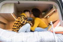 Side view of diverse man and woman kissing each other while sitting on bed in modern van during road trip — Stock Photo