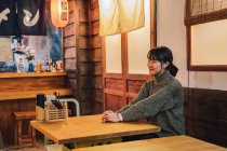 Content Asian woman in casual sweater looking away with toothy smile while sitting at wooden table in ramen bar — Stock Photo