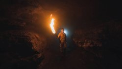 Joven espeleólogo masculino con antorcha encendida de pie en la oscura cueva rocosa estrecha mientras explora el entorno subterráneo mirando a la cámara - foto de stock