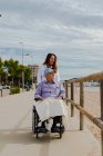 Delighted adult daughter pushing wheelchair with senior father and enjoying stroll along promenade near sea — Stock Photo