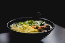 Bowl of savory Asian soup with noodles against dark background in cafe — Stock Photo