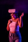 Hembra irreconocible en auriculares VR interactuando con la realidad virtual mientras está de pie en un estudio oscuro con iluminación de neón azul y rosa y vapor - foto de stock
