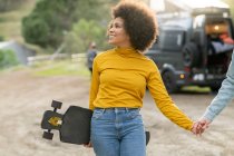 Ethnic African American young woman with longboard holding hands with cropped boyfriend while walking on countryside road on summer weekend day near caravan — Stock Photo