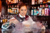Jeune femme barman qualifiée utilisant pistolet à fumée saveur bluster tout en garnissant cocktail au comptoir du bar — Photo de stock