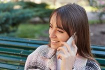 Desde arriba, una mujer milenaria sonriente moderna con un elegante atuendo de primavera sentada en el banco y respondiendo a una llamada telefónica mientras descansa en la calle urbana mirando hacia otro lado. - foto de stock