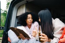 Cheerful young multiracial women drinking beer and discussing travel route with map while chilling together in camper van during summer journey — Stock Photo