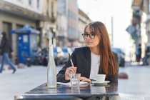 Позитивная молодая деловая леди в элегантном костюме и очках делает заметки в блокноте, сидя за столом в уличном кафе в городе, отворачиваясь — стоковое фото