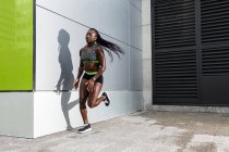 Vue latérale de l'athlète afro-américaine bondissant vers l'avant tout en courant près du mur de bâtiment moderne sur la rue de la ville — Photo de stock