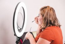 Vista lateral da fêmea gorda usando escova para aplicar maquiagem perto da luz do anel no estúdio — Fotografia de Stock