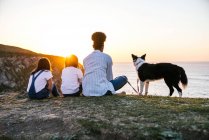 Visão traseira da mãe irreconhecível com crianças e cão Border Collie sentado na costa perto do mar e admirando o pôr do sol enquanto passam a noite juntos na praia — Fotografia de Stock
