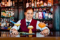 Lächelnde Barkeeperin an der Theke mit verschiedenen Arten von Alkoholgetränken, die in kreativen Cocktailgläsern in Form von Pilzen und Fisch serviert werden — Stockfoto