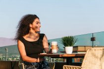 Счастливая молодая кудрявая латиноамериканка пьет кофе на вынос и наслаждается летним днем, сидя за столом на террасе кафе — стоковое фото