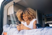 Felice femmina afroamericana con i capelli ricci sorridenti e navigando cellulare mentre riposava sul letto in roulotte durante il viaggio in auto — Foto stock