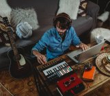 Сверху счастливый молодой человек в наушниках с помощью синтезатора и ноутбука за столом дома — стоковое фото