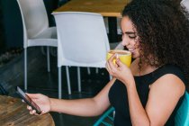Vista lateral do feliz jovem hispânico feminino navegando telefone celular enquanto desfruta de delicioso cappuccino na mesa de café de rua — Fotografia de Stock