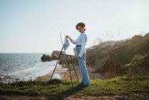 Вид сбоку на молодую женщину в стильной одежде и берете, стоящую на травянистом побережье возле песка и океана в солнечный день, рисуя картину кистью на холсте на мольберте — стоковое фото