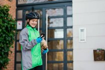 Livraison heureuse femme portant des boîtes enveloppées et naviguant sur la carte GPS sur le téléphone mobile tout en se tenant sur la rue résidentielle le jour ensoleillé — Photo de stock